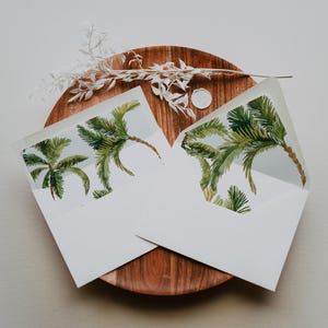 May include: Two white envelopes with a tropical palm tree design on the flap. The envelopes are on a wooden plate with a sprig of dried white flowers.