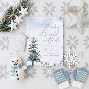 Op de afbeelding: Een rechthoekige uitnodigingskaart voor een babyshower met een aquarelillustratie van een besneeuwde dennenboom, blauwe sneeuwvlokken en de tekst Baby it's cold outside, Ethan Miles, Saturday, 09.12.2023, Miles Family House, 4530 Glensythe Road, Brenham, TX 77833.