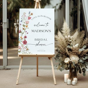 Editable “Love is in Bloom” bridal shower welcome sign template featuring a vertical border of soft watercolor wildflowers in pink, burgundy, lavender, and yellow, with modern script and serif fonts on a minimalist white background