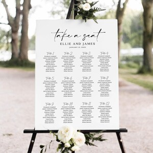 May include: A white wedding seating chart on a black easel. The chart reads "take a seat" above the names "Ellie and James" and the date "January 15, 2023." Twelve tables are listed with guest names. Floral arrangements adorn the easel.