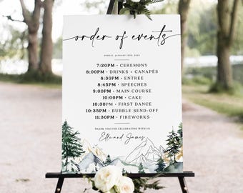 May include: A white wedding sign on a black easel displays the "order of events" in elegant script. The schedule includes ceremony, drinks, speeches, and fireworks, with a mountain and tree illustration at the bottom. The sign is decorated with white flowers.