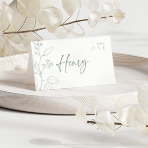 Puede incluir: Una tarjeta de mesa blanca con el nombre "Henry" en letra cursiva. La tarjeta tiene un diseño floral y las palabras "Table One". Está sobre una bandeja de madera, decorada con hojas.
