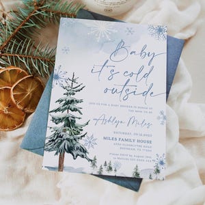 “Baby It’s Cold Outside” baby shower invitation with watercolor pine trees, scattered snowflakes, and elegant script on a soft winter background.