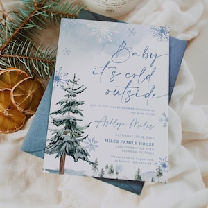May include: A baby shower invitation with a winter theme. The invitation features the text "Baby it's cold outside" in elegant script, along with event details. The design includes watercolor snowflakes and a snowy pine tree illustration, set against a blue background.