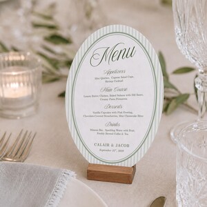 May include: An oval-shaped menu card with a sage green and white vertical striped border, featuring the text Menu, Appetizers, Main Course, Desserts, Drinks, Calair & Jacob, and September 21st, 2029, held upright by a small rectangular wooden block stand.
