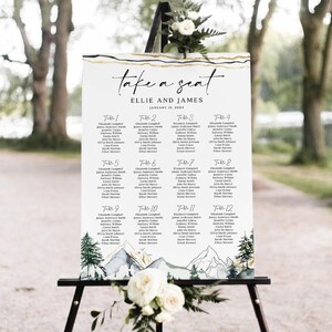 May include: A white wedding seating chart with gold accents and a watercolor mountain scene. The sign reads "take a seat" and lists names under table numbers. The date is January 15, 2023. The sign is displayed on a black easel.