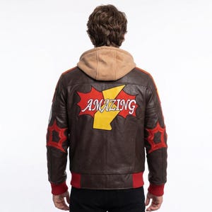 May include: A brown leather jacket featuring a tan hood and a large red and yellow graphic on the back with the word AMAZING in white text. The jacket includes red star-shaped accents on the sleeves and a red ribbed hem at the waist and cuffs.