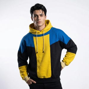 Könnte beinhalten: Ein farbblockierter Kapuzenpullover in Schwarz, Blau und Gelb. Der Kapuzenpullover hat eine gelbe Kapuze und ein vertikales gelbes Panel auf der Vorderseite. Die Ärmel sind schwarz mit gelben Bündchen. Der obere Teil ist blau über den Schultern. Der Kapuzenpullover hat einen Kordelzug.