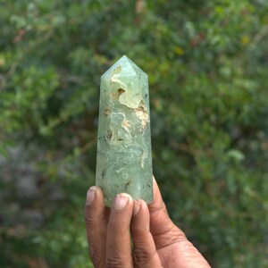 Pode incluir: Uma ponta de cristal verde claro com um topo facetado. O cristal tem inclusões e uma textura ligeiramente áspera. O cristal é segurado em uma mão contra um fundo verde desfocado.
