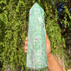 Puede incluir: Un alto obelisco de cristal verde con una parte superior puntiaguda. El obelisco está salpicado de pequeñas inclusiones redondas de color magenta. El cristal se sostiene en la mano de una persona contra un fondo de follaje verde.