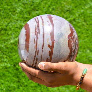 May include: A polished stone sphere with a mix of white and reddish-brown streaks. The sphere is held in cupped hands against a backdrop of green grass. A bracelet with green and brown beads is visible on the wrist.