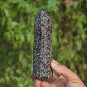 Può includere: Un alto obelisco di cristallo rettangolare con la parte superiore a punta. La pietra è un misto di grigio scuro e nero con grandi macchie irregolari di rosa intenso. L'obelisco è tenuto in mano su uno sfondo verde sfocato.
