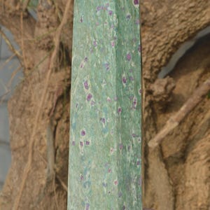 Może przedstawiać: Wysoki, prostokątny zielony obelisk kryształowy ze spiczastym wierzchołkiem. Kamień jest nakrapiany małymi fioletowymi i niebieskimi wtrąceniami. Obelisk trzymany jest w dłoni, z rozmytym pniem drzewa w tle.