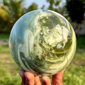 Puede incluir: Una piedra esférica pulida que presenta un patrón jaspeado de remolinos en verde bosque profundo, salvia y blanco crema. La superficie es lisa y reflectante, mostrando variaciones minerales naturales y líneas orgánicas fluidas.