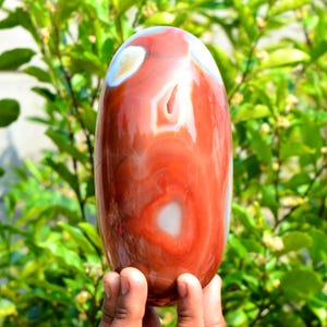Könnte beinhalten: Ein polierter, oval geformter Karneol-Achatstein mit einer glatten, glänzenden Oberfläche, die wirbelnde Muster in tiefem Rot-Orange, gebranntem Orange und cremeweißen Einschlüssen sowie eine kleine, natürlich entstandene Kristallhöhle aufweist.