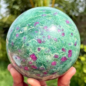 Pode incluir: Uma pedra esférica polida com uma base de cor verde vibrante e numerosas inclusões cristalinas magenta e rosa escuro espalhadas pela superfície, criando um padrão natural manchado em todo o objeto mineral redondo.