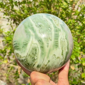 Puede incluir: Una piedra preciosa esférica pulida con un patrón arremolinado de verde y blanco. El orbe se sostiene en una mano, con un telón de fondo de follaje verde. La superficie de la piedra tiene una apariencia veteada.