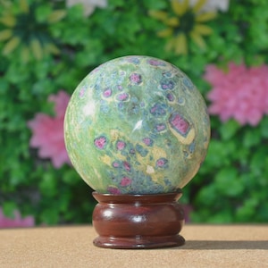 May include: A green and pink orb, possibly a gemstone, sits on a dark brown wooden stand. The orb has a speckled pattern of pink and blue against a green background. The stand is round with a flared base. The background is a blurred mix of green and pink.