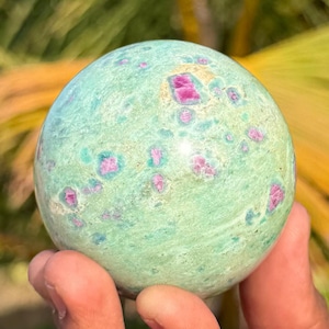 May include: A polished spherical stone featuring a light mint green base colour with scattered, irregular magenta and deep pink inclusions throughout the surface. The sphere displays a smooth, rounded texture with natural mineral variations.