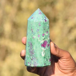 Puede incluir: Una torre de cristal de rubí zoisita verde y rosa. La piedra pulida tiene forma rectangular con la parte superior puntiaguda. El color base verde está salpicado de vibrantes manchas rosas y moradas.