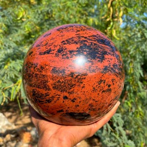 Könnte beinhalten: Eine polierte Kugel aus Mahagoni-Obsidian, mit einem satten rotbraunen Grundton und auffälligen schwarzen Mustern. Der Orb ist glatt und reflektierend und zeigt natürliche Farb- und Texturvariationen. Ein dekoratives und einzigartiges Objekt.