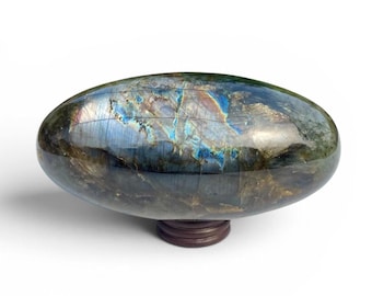 May include: A polished, oval-shaped labradorite stone featuring deep blue, grey, and iridescent flashes across its surface. The stone is mounted on a small, circular, dark brown wooden base, providing a stable display for the mineral specimen.