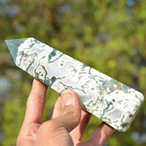 May include: A polished, rectangular gemstone with a pointed top. The stone features a white base with intricate patterns of green and brown, creating a natural, organic appearance. The gemstone is held in a hand, showcasing its size and detail.