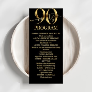 May include: A black 90th birthday program with gold lettering, detailing the event schedule. The program is placed on a white, wooden plate, offering a contrast in textures and colours. The text includes the word "PROGRAM" and the number "90".