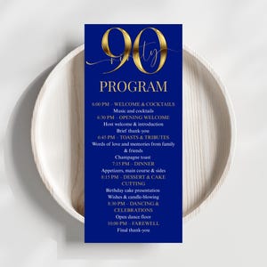 May include: A navy blue 90th birthday program card with gold script. The program includes the schedule for the event, such as welcome, toasts, dinner, and dancing. The card is set on a white wooden plate.