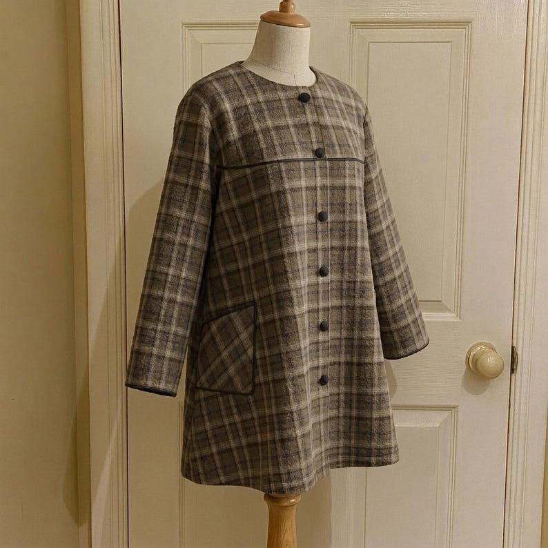 May include: A long-sleeved coat featuring a brown and grey plaid pattern, a round neckline, and a front button closure. The garment includes a horizontal seam across the chest and a single patch pocket on the lower right side with dark trim.
