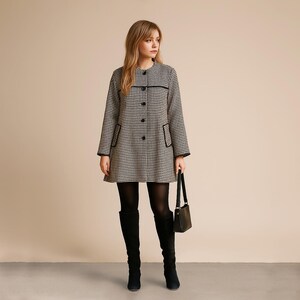 May include: A houndstooth patterned coat featuring a round neckline, long sleeves, and a front button closure. The coat includes two front pockets with black trim, matching the black piping detail across the chest and along the sleeve cuffs.