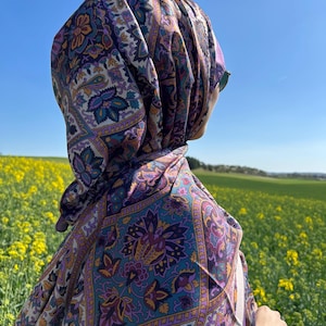 May include: A woman wearing a purple and white patterned headscarf with a paisley design. The scarf is tied around her head and neck, and she is looking off to the side. She is standing in a field of yellow flowers.