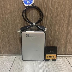 May include: A silver WD My Passport external hard drive with a black USB cable and an 8MB memory card. The memory card has the text "FMCB" and "8MB Memory Card" printed on it. The USB cable has a sticker attached.