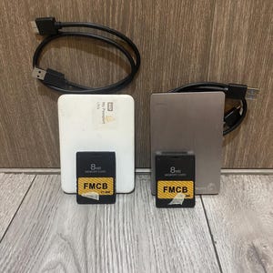 May include: Two external hard drives, one white and one silver, each paired with a black USB cable. Two black 8MB memory cards are positioned in front of the drives, each featuring a yellow label with the text FMCB V1.966 printed in black.