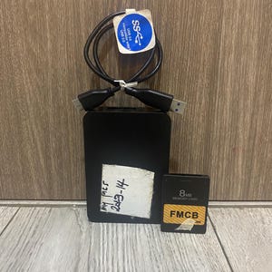 May include: Black external hard drive with a USB cable and a small 8MB memory card. The hard drive has a white label with handwritten text. The memory card has a gold and black label with the text "FMCB".