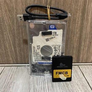 May include: A 1TB WD Blue hard drive in a clear plastic case with a black USB cable and a black 8MB FMCB memory card. The hard drive label displays technical specifications, a barcode, and the model number WD10SPZX. The memory card features text reading 8MB MEMORY CARD and FMCB V1.966.