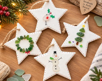 May include: Four white ceramic star ornaments with jute twine hangers. Designs include a robin with holly, a green sea glass wreath, a snowman made of sea glass, and a sea glass Christmas tree. Each star features hand-painted red and green dots.