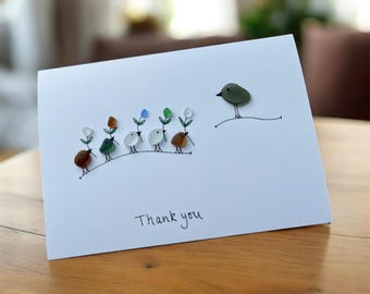 May include: A white greeting card with a hand-drawn design of birds and flowers made from colorful stones. The text "Thank you" is written in cursive at the bottom. The card is on a wooden surface.