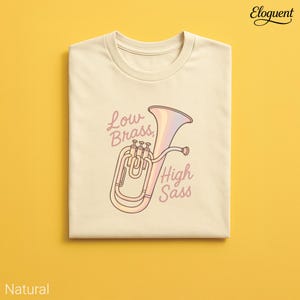 Könnte beinhalten: Ein gefaltetes, naturfarbenes T-Shirt mit einer Cartoon-Illustration eines Euphoniums in Rosa- und Lilatönen. Der Text "Low Brass / High Sass" ist in Schreibschrift geschrieben. Das Shirt befindet sich auf einem gelben Hintergrund.