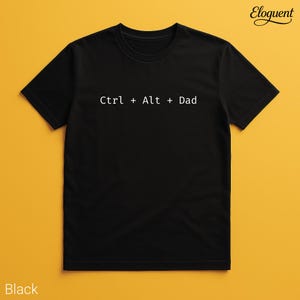Op de afbeelding: Zwart T-shirt met ronde hals en de tekst "Ctrl + Alt + Dad" in het wit. Het T-shirt is effen zwart en heeft korte mouwen. Het T-shirt wordt gepresenteerd op een gele achtergrond.