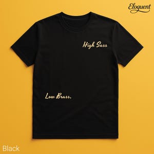 Puede incluir: Una camiseta negra de manga corta con el texto High Sass en letra dorada en la parte superior derecha del pecho y el texto Low Brass, en letra dorada en la parte inferior izquierda. La prenda tiene cuello redondo y corte estándar.