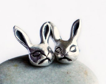 May include: Two silver-colored rabbit head sculptures with closed eyes and downturned mouths rest on a smooth, gray stone. The sculptures have detailed features, including long ears and textured fur. The background is a plain white.