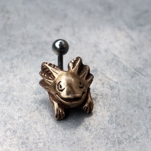Puede incluir: Un amuleto de axolotl de color dorado con un poste plateado, perfecto para la elaboración de joyas o para agregar un toque único a un proyecto.