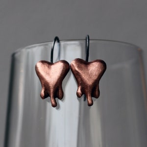 Könnte beinhalten: Ein Paar Ohrringe mit einem tropfenden Herzdesign. Die Herzen sind aus Kupfer und haben eine warme, rotbraune Farbe. Die Ohrringe hängen an schwarzen Metallhaken.