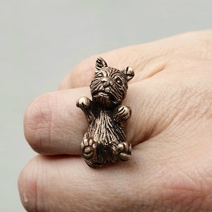 May include: A bronze ring featuring a detailed sculpture of a small dog sitting up with its paws raised. The ring is worn on a person's finger.
