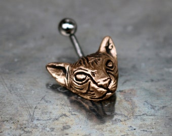 May include: A gold-colored cat head body piercing with a silver ball on the top.