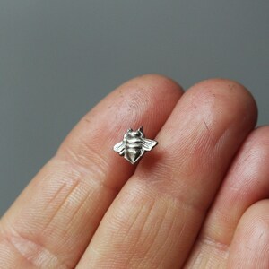 Può includere: Un ciondolo a forma di ape in argento, mostrato su un dito.