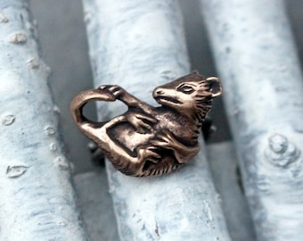 May include: A bronze ring featuring a detailed sculpture of a possum. The possum is curled up in a playful pose, with its tail wrapped around its body. The ring is set against a backdrop of white birch bark.