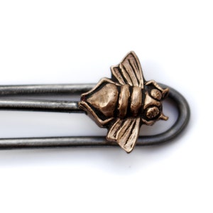 May include: A silver metal hair clip with a bronze bee charm. The bee is detailed with wings, legs, and antennae.