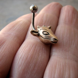 Könnte beinhalten: Ein goldfarbener Ziegenkopf-Anhänger, der an einer silbernen Körperpiercingstange befestigt ist.